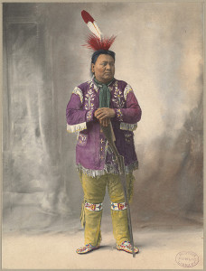 Colorized photograph of a Native American man, standing, propping his hands on a rifle that is pointed towards the floor.  He wears a large red and white feather in his hair, a purple shirt with white fringe, yellow pants with a beaded design on the knees, and beaded moccasins.
