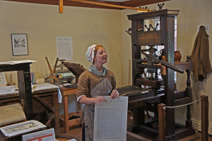 Photo of a woman in period costume and bonnet standing in front of an antique printing press, holding a portfolio-sized page of the Declaration of Independence