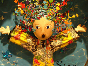 Photo of a wooden doll with mouth open and arms extended in a singing pose.  The doll has brightly colored wire forms for hair and hand-drawn patterns on its shirt