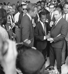 Black and white photo showing James Baldwin, a short man in a suit with a big smile, standing between Charlton Heston and Marlon Brando. A large crowd surrounds them.