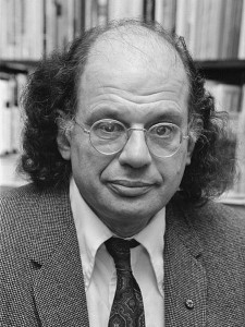 Black and white photo of Ginsberg, against a backdrop of books.  He is bald on top with shoulder-length hair on the sides, and wears wire-framed glasses and a suit.