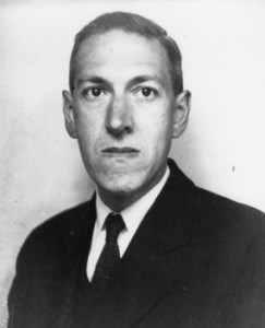 Black and white photo of Lovecraft.  He has a stern expression as he stares into the camera.  He wears a dark suit and is standing against a plain white background.