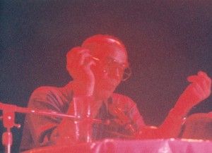Low-quality photo showing Thompson seated at a table in front of a microphone.  His figure appears in red tones, and the background is purple.