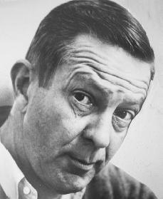 Black and white candid photo of John Cheever.  He is looking up at the camera with a raised eyebrow.