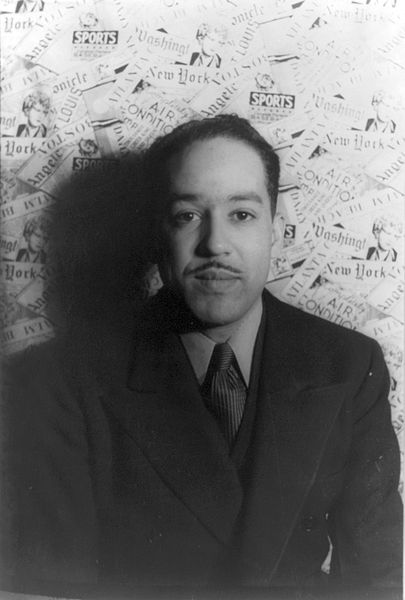 Black and white photo of Hughes, who is standing against a wall covered in newspaper-styled wallpaper.  He wears a suit and tie, has a thin mustache, and is staring directly into the camera.