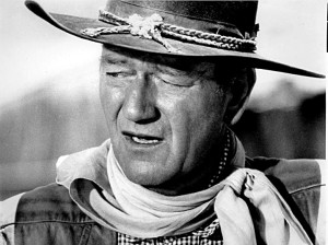 Black and white photo of John Wayne, as a photo still from a western movie.  He is wearing a cowboy hat, a scarf tied around his neck, and a plaid shirt.  He is squinting and looking off to the left, with his mouth partially open as if speaking.