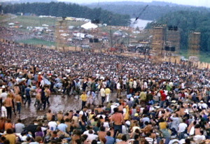 Photo of a crowd scene on a hillside.  A path through the middle of the crowd is very muddy.  A stage appears at the bottom of the hill, with a lake and woods behind it. 