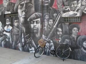 Photo of a wall mural depicting figures from the Black Panther movement, many African-American men with Afros or wearing berets.  The central figure looks into the street and holds a machine gun.  A bicycle is parked in front of the mural.