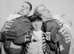 Black and white photo of The Three Stooges.  All are dressed in comedic military costumes with ruffles and buttons on their jackets.  Curle and Larry are squeezing against Moe, squished in the middle with a pained expression on his face.