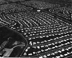 Black and white aerial photo showing hundreds of houses in a development, roughly centered around a larger white structure, possibly a school or church
