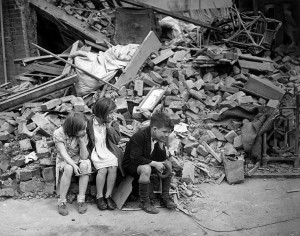 Black and white photo of three children sitting at the edge of a pile of rubble.  They all have their heads propped on their hands.  Furniture and other household goods are evident in the rubble.