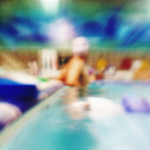 Very blurry photo of a person in a bathing cap standing in a pool.  Shapes are distorted, and colors become prominent: the aqua of the water, a blue towel on the side of the pool, the white bather's cap, and the dark blue wall above the pool.