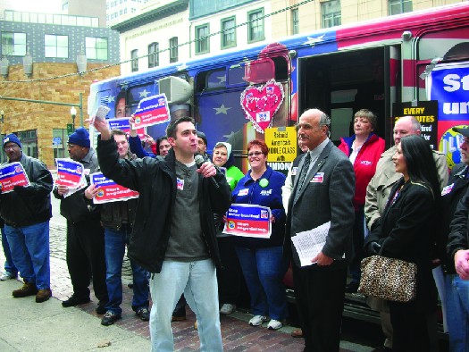 An image of a group of people standing in front of a bus. Some of the people hold signs.