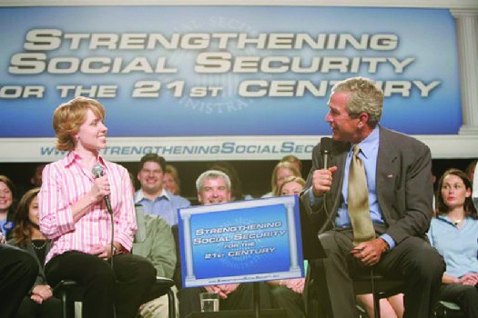 A photo of George W. Bush speaking at an event. The banner behind him says 