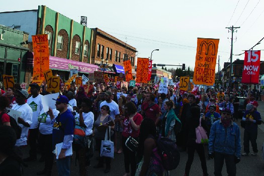 An image of a group of people marching down a street, one of whom holds a sign that reads 