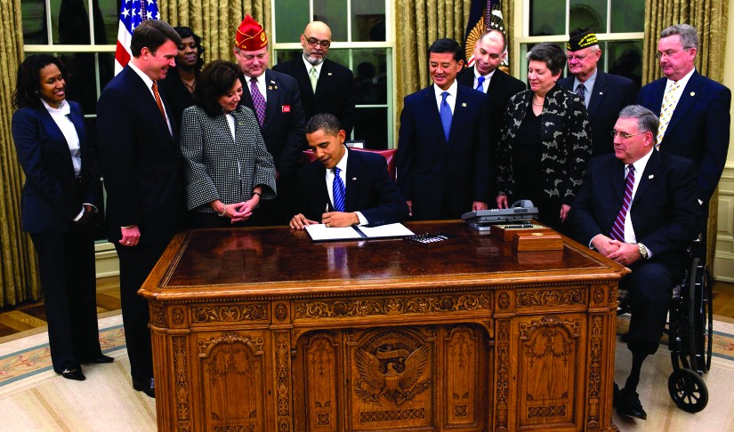 An image of a group of 12 people standing around Barack Obama, who is seated at a desk and signing a piece of paper.