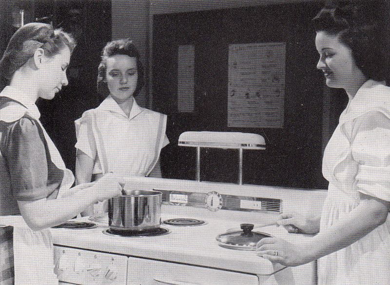 Three women gather around a stovetop in a Home Economics class.