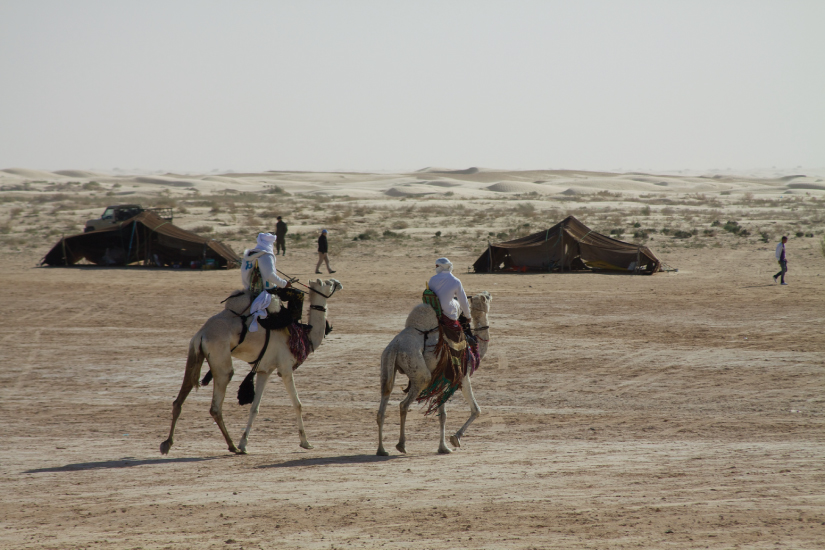 This photo shows two white-clad men riding camels through a sparse desert. Two canvas tents are visible in the background.