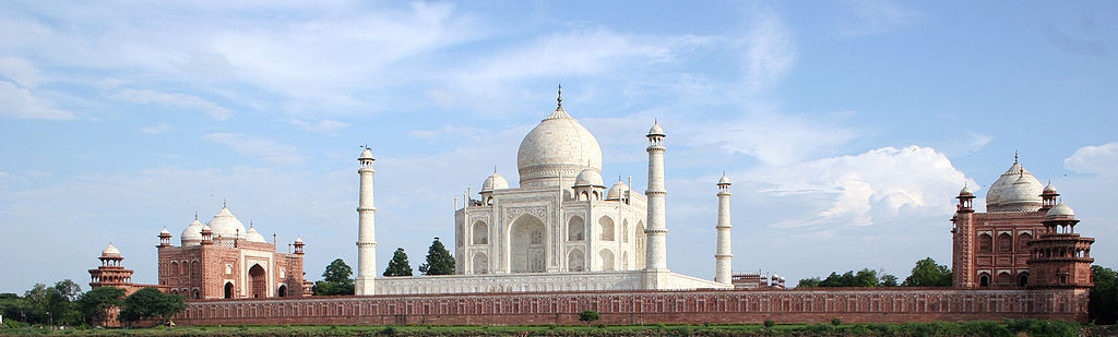 Photograph showing the entire complex of the Taj Mahal. The complex is made of three buildings. The center building is the most grand and pure white, while the other two are smaller and use red-brown brick along with white. The central building is framed by four domed towers atop a shorter fence: these are all white. The central building has a large dome in its center, and four domes on its corners. The front has a large archway leading to its entrance, and there are arched balconies on the ground floor as well as the second floor. Even at a distance, there is evidence of intricate carvings, especially on the entrance.