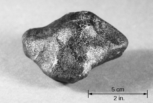 Photograph of a Piece of Vesta. This photo shows an irregularly shaped metallic fragment from Vesta. The scale at lower right reads