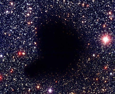 Photograph of Barnard 68. An absolutely black, comma-shaped cloud is centered in this image, with hundreds of stars seen in the background.
