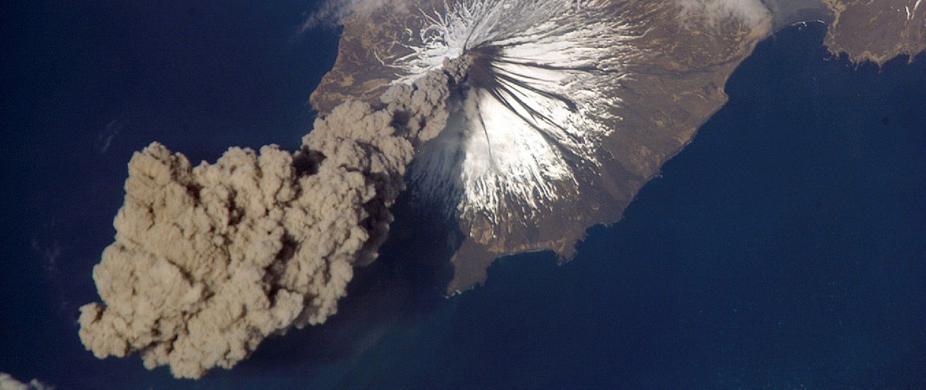 Image of a Volcanic Eruption Taken from Space. A huge plume of dark grey smoke emerges from the snow-covered peak of the Cleveland Volcano in Alaska.
