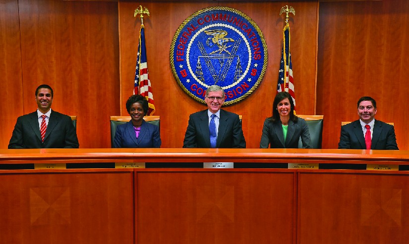 An image from left to right of Ajit Pai, Mignon Clyburn, Chairman Tom Wheeler, Jessica Rosenworcel and Michael O’Rielly seated in front of a large circular banner reading 