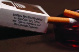 A photo of a cigarette box and two cigarettes. The cigarettes are resting in an ashtray. Text on the cigarette box reads Surgeon General's Warning. Smoking causes lung cancer, heart disease, emphysema, and may complicate pregnancy.