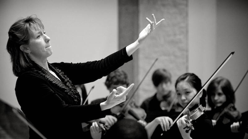 Black-and-white photo of a female conductor leading an orchestra of young musicians. Violinists pictured.