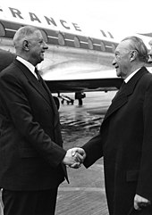 De Gualle and Konrad Adenauer shake hands in front of a plane. 