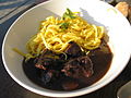 An image of boeuf bourguignon in a white bowl, which has a marinated meat and noodles on the side. 