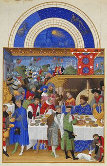 John, the Duke of Berry, is sitting with a cardinal at a high table, in front of a fireplace, tended by many servants at his dinner table.
