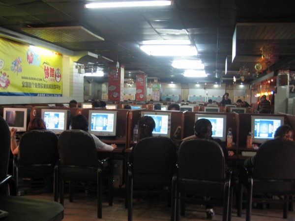 Many people sitting in chairs are shown staring at computer screens in a restaurant/café setting. Chinese posters can also be seen.