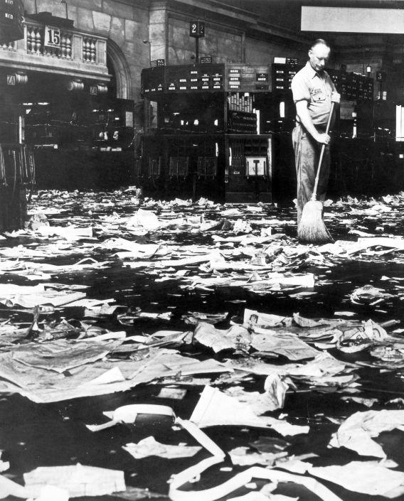A man begins to sweep the floor of a large room littered with paper trash.