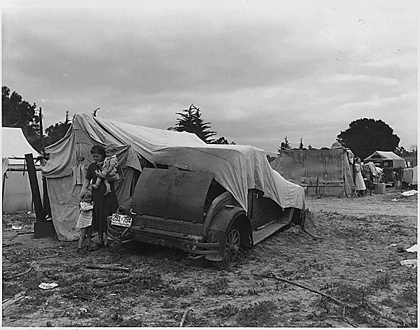 A woman stands next to an old car being used as part of a tent. The woman holds one child and has her hand on the shoulder of another young one standing by her side. Similar tents are in the background.
