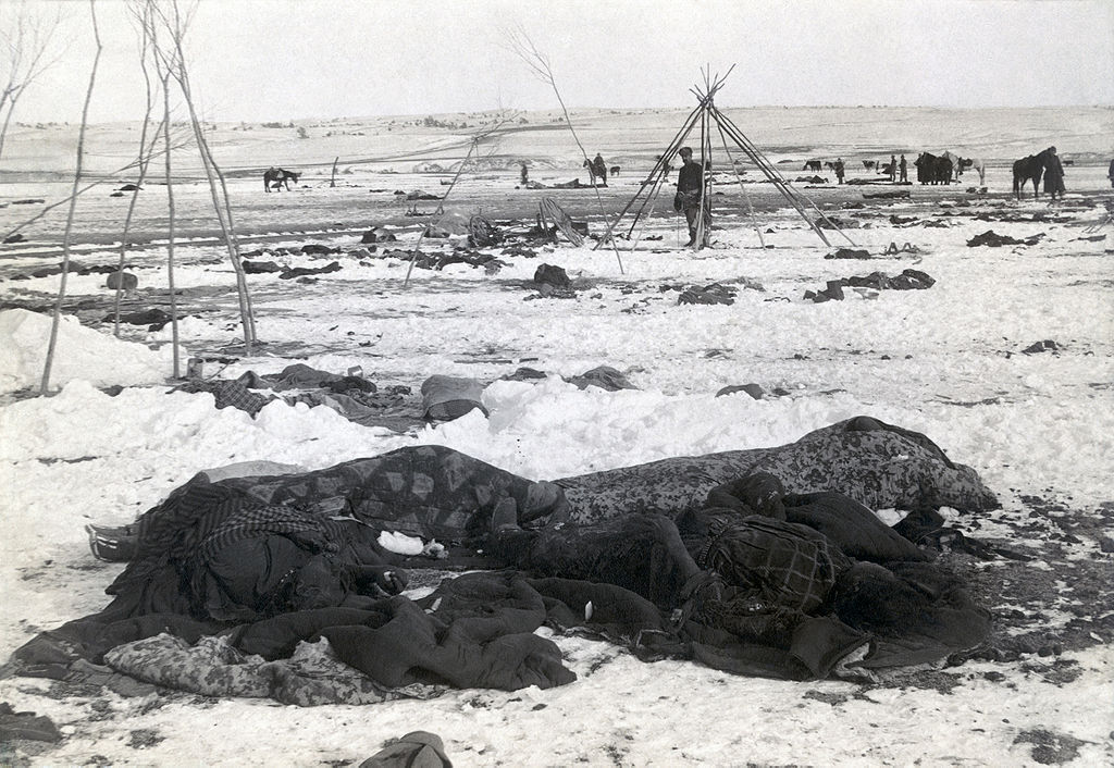 A winter scene on the prairie. Four shapes wrapped in blankets are in the foreground. Soldiers and horses are standing in the background. One man stands inside teepee poles and looks toward the camera.