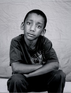 homeless boy looking reflectively at the camera for his picture, while he sits with his arms crossed in front of him on his knees.