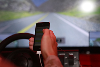 A person’s right hand is holding a cellular phone. The person is in the driver’s seat of an automobile while on the road.