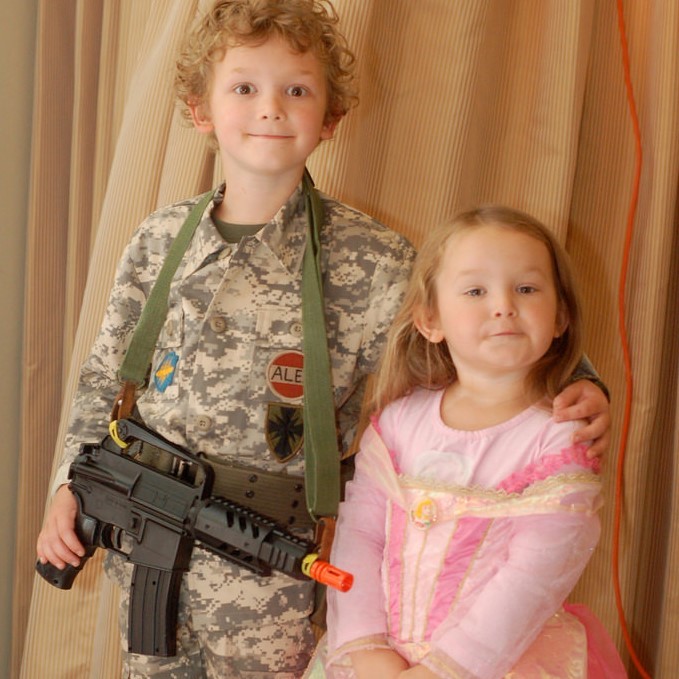 A brother and sister stand side by side. He is dressed in a camouflage military uniform and is holding a toy gun. She is dressed in a pretty pink princess dress.