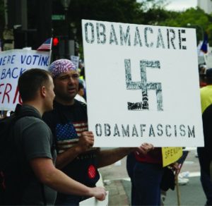 An image of a person holding a sign that reads Obamacare Obama fascism, and a Nazi swastika