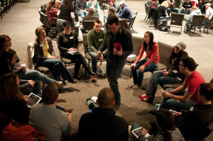 Photo of a group of young adults sitting in a ring of chairs. A man stands in the middle holding a ball of yarn.