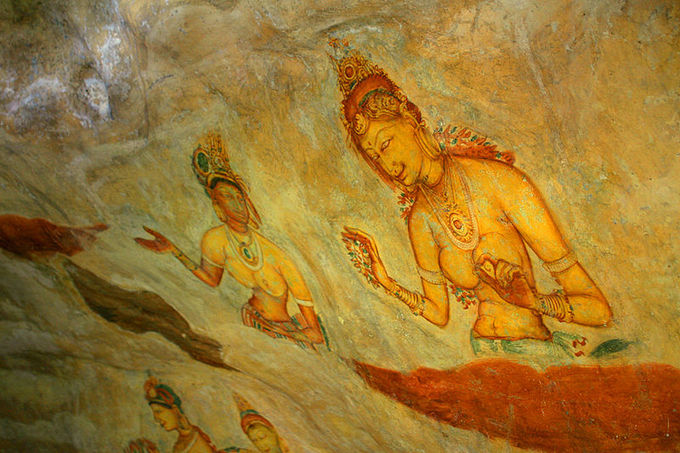 This photo shows part of the Sigiriya frescoes. It depicts topless women wearing ornate jewelry with flowers. It features bright, vibrant shades of orange, red, and yellow.
