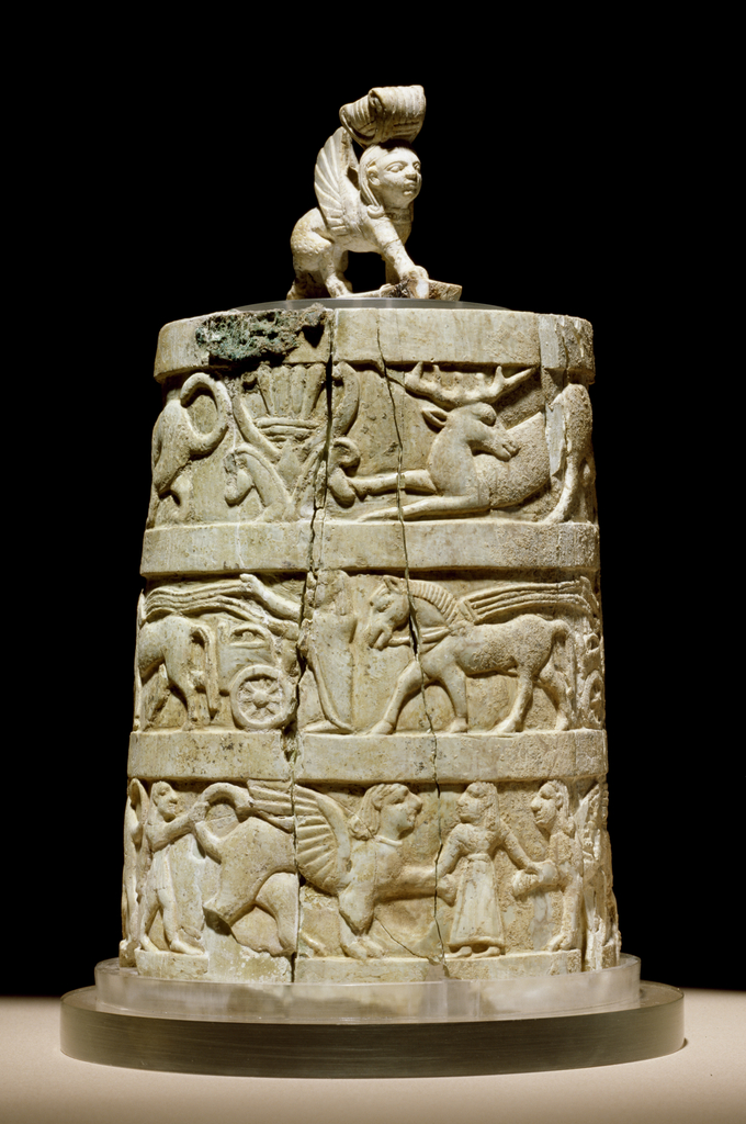 This is a photo of a pyxis (a type of box) decorated with reliefs of sphinxes, a lotus plant, and chariots. The handle of the lid takes the form of a standing sphinx wearing a lotus crown.