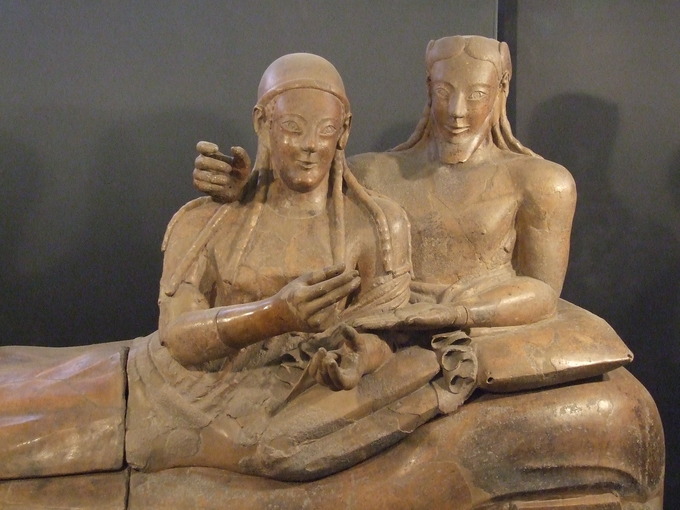 This sarcophagus depicts a man and woman sharing a banquet couch. They are both smiling and expressing affection. She is in gesture of offering something to him and he is making a gesture of receiving it. They both have almond-shaped eyes and long braided hair.