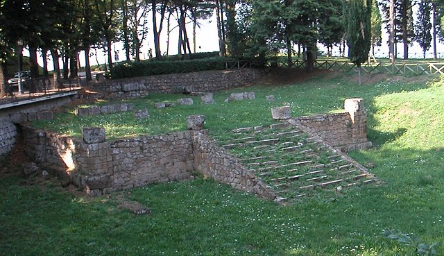 This is a photo of the ruins of the foundation and stairway of an Etruscan temple. Grass has grown over the ruins which appear to be made of stone.