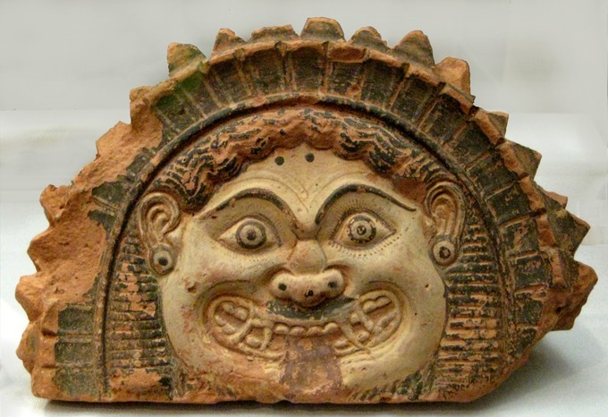 This is a photo of an antefix with the head of a Gorgon, depicted as a monstrous creature with wide eyes and a fierce grin.
