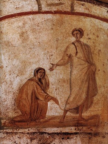 In this image, Jesus heals a bleeding woman. He stands above her wearing white robes and places a hand above her head. She kneels, appearing either to reach for him or to hold onto the hem of his robe.