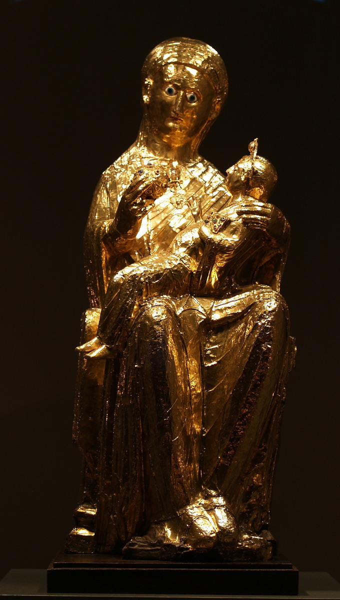 This is photo of the Golden Madonna of Essen, a sculpture of the Virgin Mary and the infant Jesus. Mary is depicted sitting on a stool, with a slightly oversized Christ child figure sitting on her lap. She wears a robe and veil. In her right hand she holds a globe with her thumb and two fingers, while her left hand supports the infant in her lap.