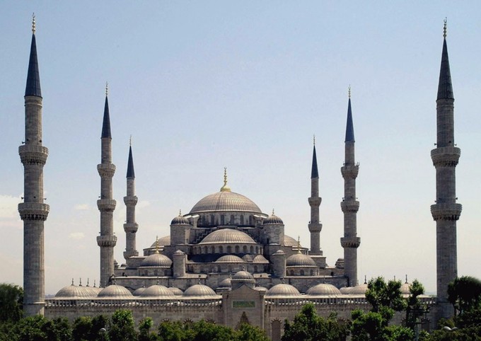 This is a photo of the Blue Mosque. In the center is a large dome, beneath are several smaller domes. All together, they form a triangular or pyramid shape. There are three slender minarets on either side of the domes.