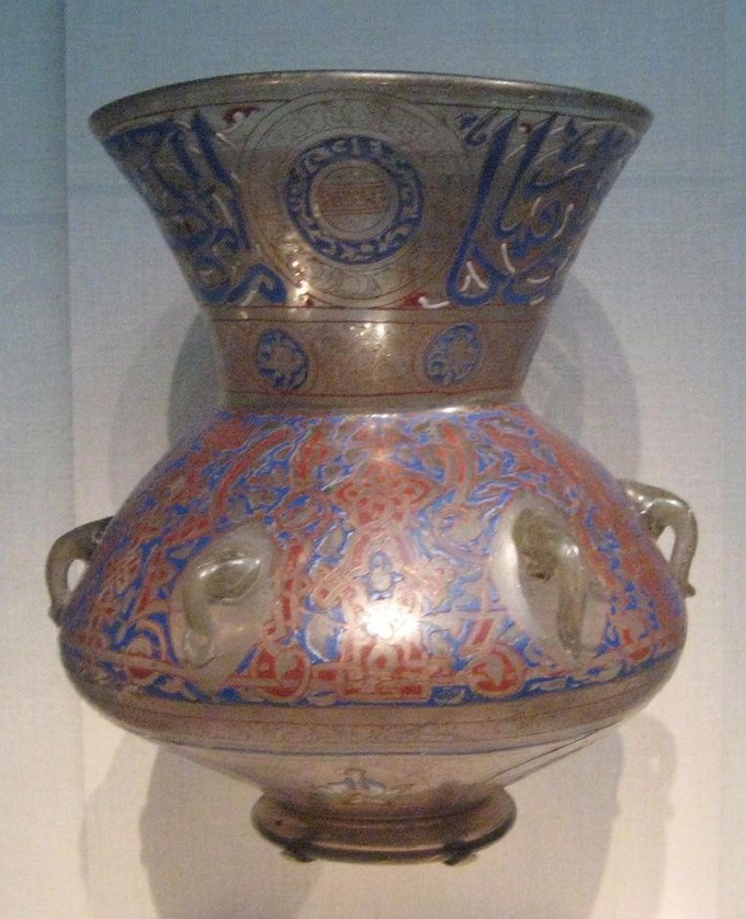 This is a photo of a glass mosque lamp, which has a large round bulbous body rising to a narrower waist, above which the top section is flared. It is bronze-colored decorated with red and blue arabesques.